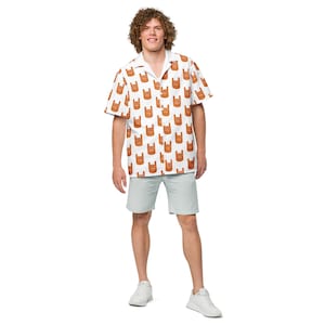 May include: A white short-sleeved button-down shirt with a brown repeated pattern of a hand making the rock and roll sign. The model is wearing light blue shorts and white sneakers.