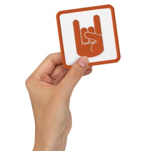 Könnte beinhalten: Ein weißer Patch mit einer orangefarbenen gestickten Hand, die die Rock'n'Roll-Handgeste macht. Der Patch ist von einem orangefarbenen Rand umgeben.