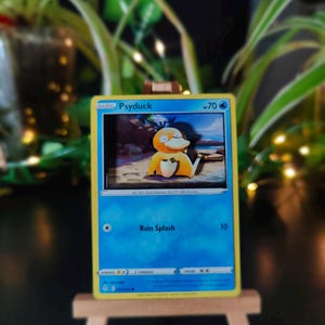 Può includere: Una carta collezionabile di Pokémon con Psyduck, una creatura gialla a forma di anatra. La carta ha uno sfondo blu con il testo "Rain Splash" e altri dettagli del gioco. È esposta su un piccolo cavalletto di legno.