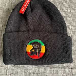 Op de afbeelding: Zwarte beanie met een rood, geel en groen geborduurd logo van een Spartaanse helm. Het logo is omgeven door een gele cirkel met een zwarte omtrek. De beanie heeft een label met de tekst "Resolute Headwear".