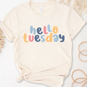May include: A cream colored t-shirt with the text "hello tuesday" in colorful letters. The letters are in shades of blue, pink, yellow, and orange.