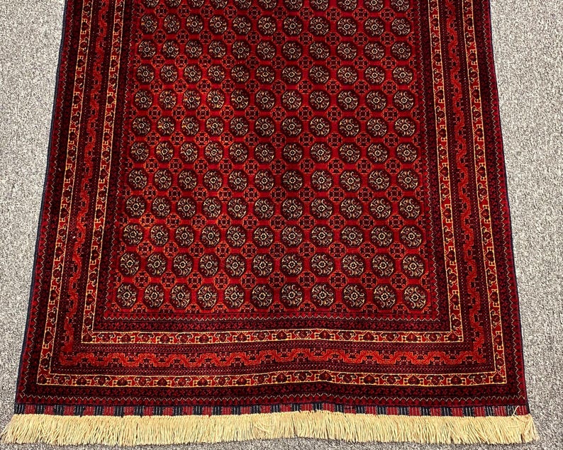 May include: A rectangular, red rug with a repeating circular pattern in shades of brown and gold. The rug has a detailed border with geometric designs and a cream-colored fringe along the bottom edge. The rug is likely made of wool.