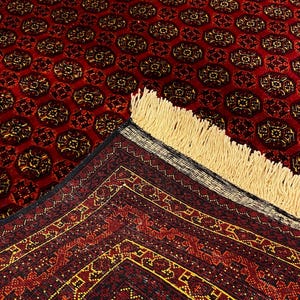 May include: A red oriental rug with a repeating pattern of dark brown and gold medallions. The rug has a detailed border with geometric designs in red, gold, and black. The fringe is a light beige color.