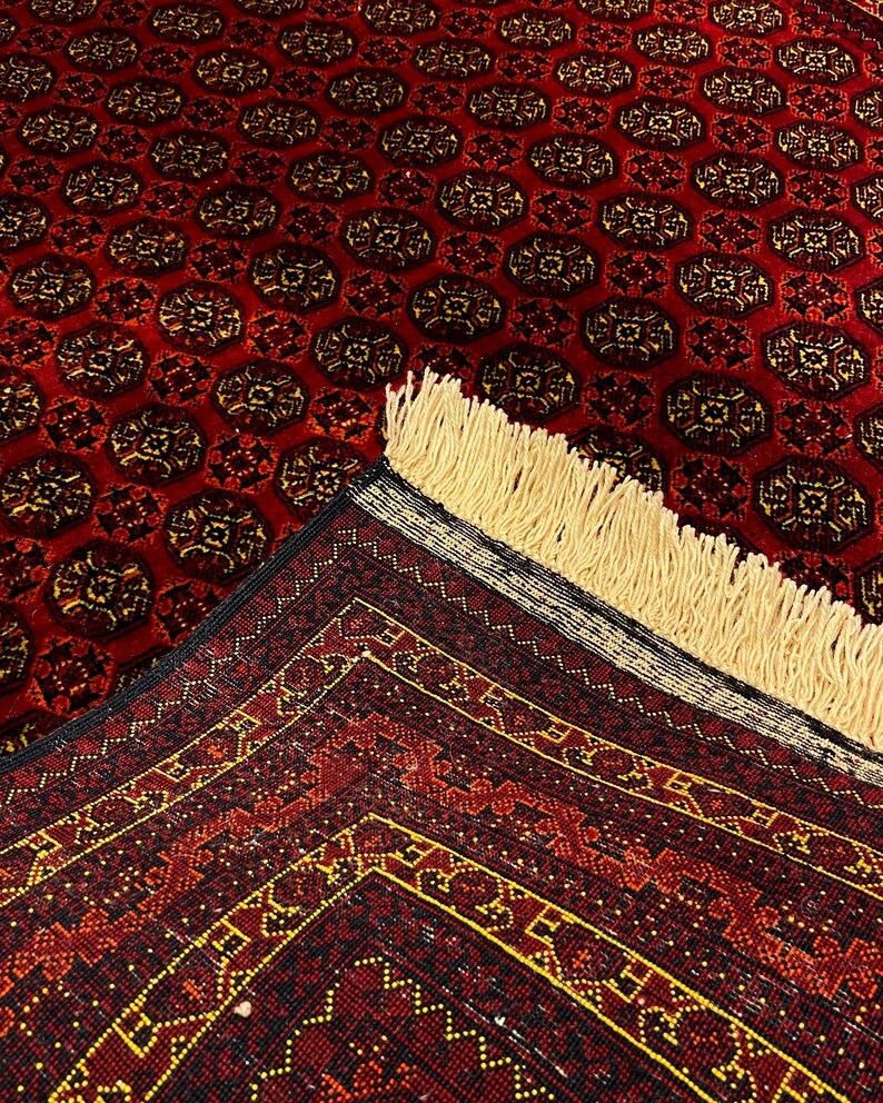 May include: A red oriental rug with a repeating pattern of dark brown and gold medallions. The rug has a detailed border with geometric designs in red, gold, and black. The fringe is a light beige color.