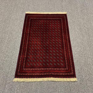 May include: A rectangular, red rug with a repeating pattern of small, stylized floral motifs. The rug has a dark border and cream-colored fringe along the short edges. The pattern is in shades of red, brown, and black.
