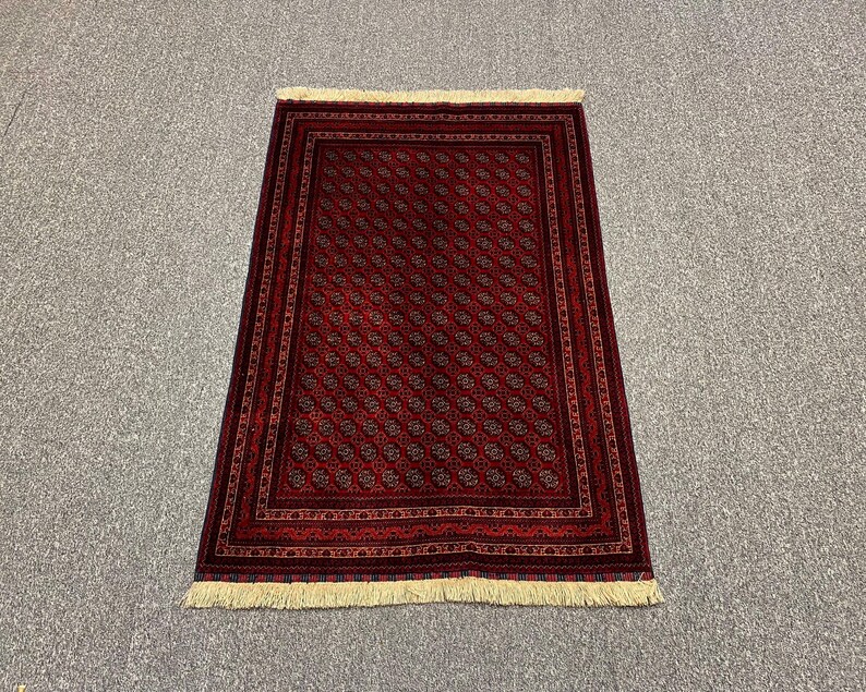 May include: A rectangular, red rug with a repeating pattern of small, stylized floral motifs. The rug has a dark border and cream-colored fringe along the short edges. The pattern is in shades of red, brown, and black.