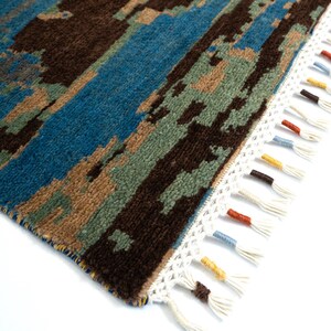 May include: A section of a rectangular rug with a pixelated design in shades of brown, blue, green, and tan. The rug has a white fringe with colorful tassels. The rug's design has a textured appearance.