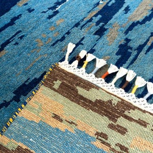 May include: A close-up of a blue and brown patterned rug with white fringe. The rug has a geometric design with shades of blue, brown, and tan. The fringe is white with accents of yellow, orange, and brown.