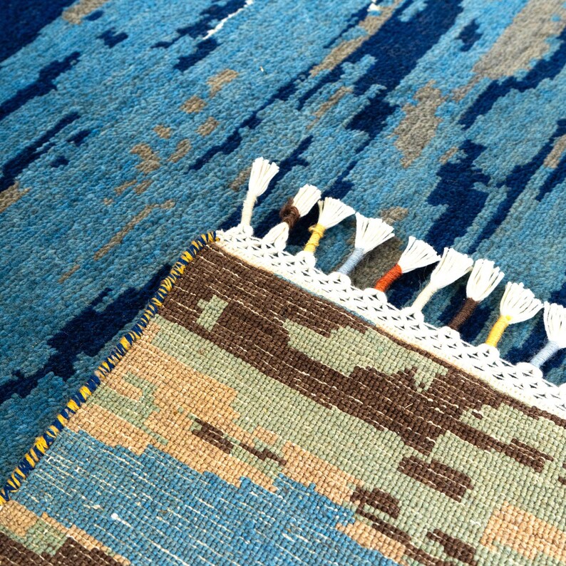 May include: A close-up of a blue and brown patterned rug with white fringe. The rug has a geometric design with shades of blue, brown, and tan. The fringe is white with accents of yellow, orange, and brown.