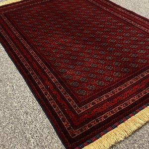 May include: A rectangular, hand-woven rug with a deep red background and intricate geometric patterns in shades of brown and gold. The rug features a detailed border and cream-colored fringe along the edges, showcasing a traditional design.