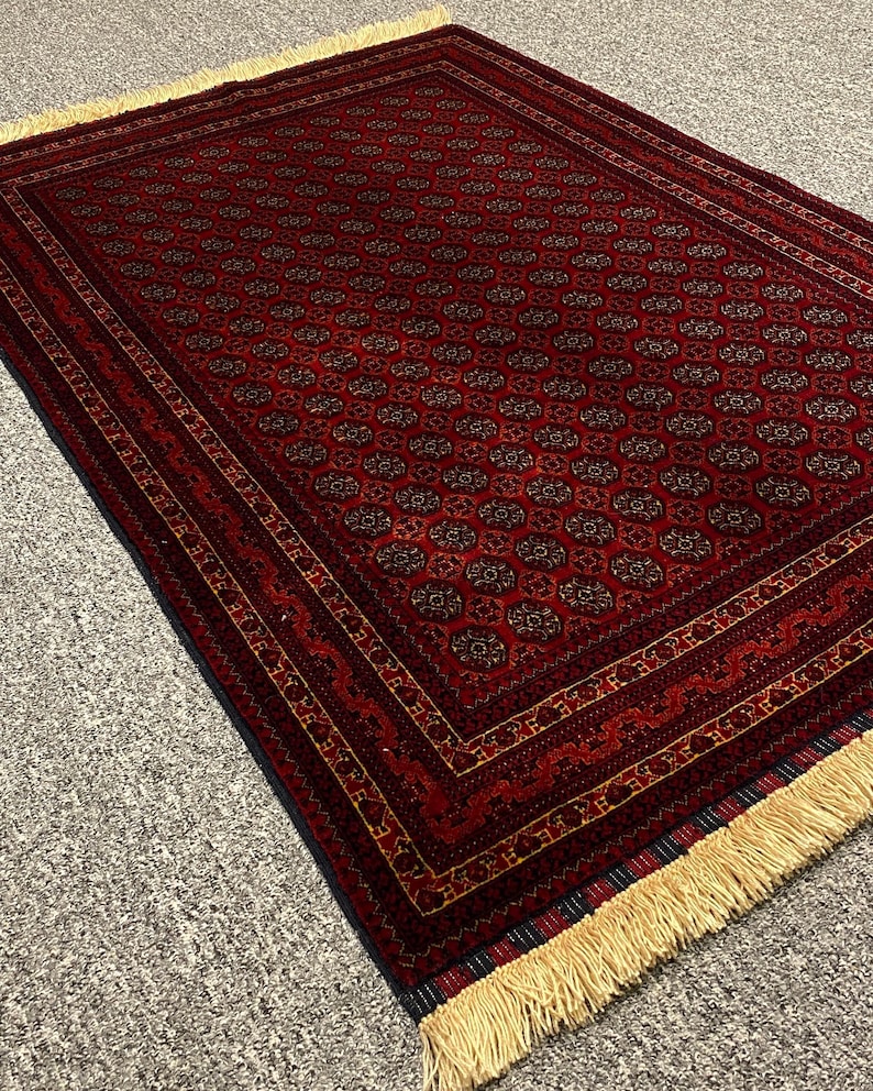 May include: A rectangular, hand-woven rug with a deep red background and intricate geometric patterns in shades of brown and gold. The rug features a detailed border and cream-colored fringe along the edges, showcasing a traditional design.