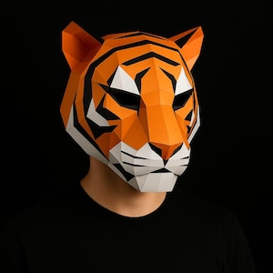 Può includere: Una maschera geometrica a forma di tigre in carta. La maschera è arancione con strisce nere e dettagli bianchi. La persona indossa una maglietta nera. Lo sfondo è nero, evidenziando i colori vivaci della maschera.
