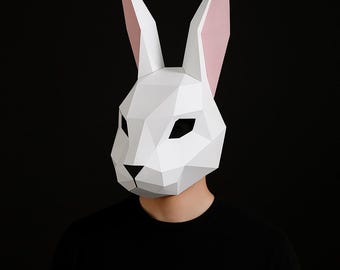May include: A white geometric rabbit mask with pink inner ears. The mask has a polygonal design and covers the face, with black eye openings and a black triangle for the nose. The person is wearing a black shirt.