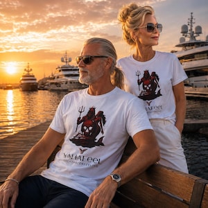 Könnte beinhalten: Ein weißes Kurzarm-T-Shirt mit einem Grafikdruck einer Figur, die einen Dreizack über einem Totenkopf hält, sowie dem Text YA HAFEEL YACHTHAFENFEELING. Das Design ist mittig auf der Brust platziert und zeigt eine dunkelrote und schwarze Illustration.