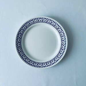 May include: A white ceramic plate with a blue and white patterned border. The pattern features a repeating design of stylized waves.