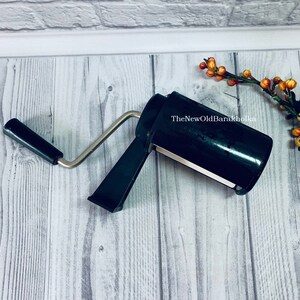 May include: A black hand-cranked pasta maker with a silver handle. The maker is on a white wood surface. The text "TheNewOldBarakholka" is on the side of the maker.
