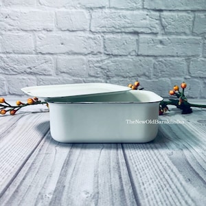 Puede incluir: Contenedor de almacenamiento rectangular de esmalte blanco con tapa. El contenedor tiene un borde negro y la tapa tiene una pequeña manija de metal.