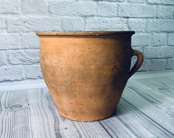 Pode incluir: Um vaso de terracota com uma única alça. O vaso é marrom e tem uma aparência rústica.