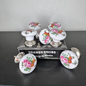 May include: A set of white ceramic drawer knobs with a floral design in pink, orange, and green. Each knob has a silver metal base and a butterfly accent. The box reads "DRAWER KNOBS".