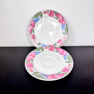 May include: Two white ceramic saucer plates with a floral design. The plates feature a pink, blue, and green floral pattern around the rim. The center of each plate is white with a recessed area for a cup.
