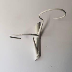 Puede incluir: Un soporte de pared de metal blanco roto para plantas. El soporte presenta una parte superior circular y un estante horizontal curvado. El diseño es minimalista e industrial, con un acabado ligeramente desgastado. El soporte está montado en una pared blanca.