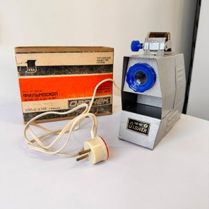 May include: A vintage film projector with a gray metal body and a blue knob. The projector is plugged into a white electrical cord. The box for the projector is labeled "Фильмоскоп" in red letters.