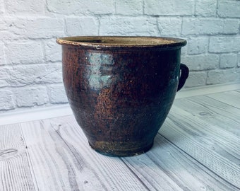 Pode incluir: Um vaso de cerâmica marrom com uma alça de um lado. O vaso tem uma superfície áspera e texturizada e é ligeiramente envidraçado.