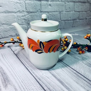 Puede incluir: Tetera blanca con ribete dorado y diseño floral naranja y negro. La tetera tiene tapa y asa. El texto "The New Old Dachakholka" es visible en el lateral de la tetera.