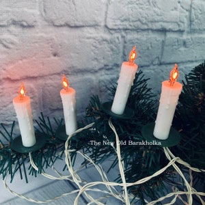 Pode incluir: Uma corda de luzes de Natal brancas em forma de vela com lâmpadas vermelhas, colocada sobre uma guirlanda de abeto artificial verde. As luzes estão ligadas e acesas, projetando um brilho quente. O texto "The New Old Barakholka" é visível na imagem.