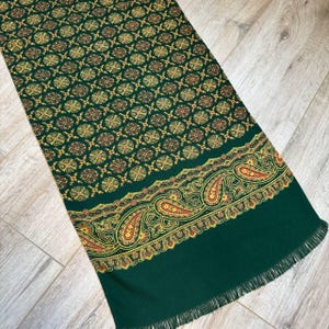 Könnte beinhalten: Ein grüner Schal mit einem sich wiederholenden Muster aus goldenen und braunen floralen Designs. Der untere Rand weist ein Paisley-Design in Rot, Gelb und Gold auf. Der Schal hat einen Fransenrand.