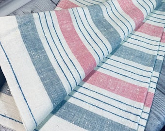 May include: A folded, striped textile in shades of white, light blue, and pink. The stripes are vertical, with thin navy blue lines accenting the design. The fabric appears to be linen or a similar woven material, suitable for a table runner or similar home decor item.