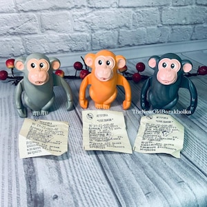 May include: Three vintage plastic monkey toys in gray, orange, and dark teal. Each monkey has a light pink face and is posed with arms outstretched. Small paper tags with Cyrillic text are in front of each toy.