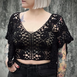Può includere: Un top in pizzo nero con un designo delicato e traforato. Il top è corto e ha una scollatura a V. La modella indossa jeans neri e ha un tatuaggio sul braccio.