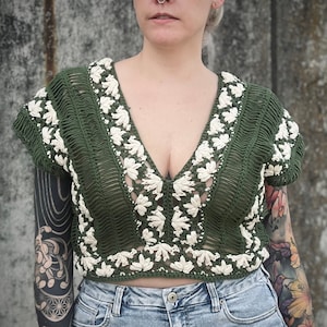 Könnte beinhalten: Ein grünes Häkeltop mit weißen Häkeldetails. Das Top hat einen tiefen V-Ausschnitt und kurze Ärmel.