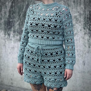 Könnte beinhalten: Ein gehäkeltes zweiteiliges Outfit in hellblau. Das Oberteil ist ein langärmliges Crop-Top mit einem zarten Spitzenmuster. Die Shorts sind hoch tailliert und haben ein ähnliches Spitzenmuster.