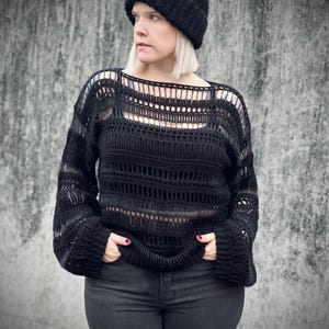 Könnte beinhalten: Ein schwarzer Häkelpullover mit lockerer Passform und einem weiten Ausschnitt. Der Pullover hat ein zartes Lochmuster und lange Ärmel. Das Model trägt den Pullover mit einer schwarzen Jeans und einer schwarzen Mütze.