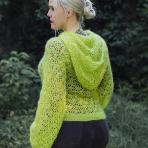 Könnte beinhalten: Ein lindgrüner, langärmeliger Kapuzenpullover mit einem komplizierten, durchbrochenen Sternen-Häkelmuster. Das Kleidungsstück hat eine flauschige, strukturierte Oberfläche, eine Kapuze und einen gerippten Saum, kombiniert mit einer dunklen Hose.