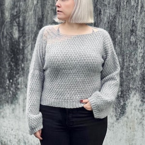 Könnte beinhalten: Ein grauer Strickpullover mit lockerer Passform und langen Ärmeln. Der Pullover hat ein strukturiertes Muster und eine leicht verkürzte Länge.