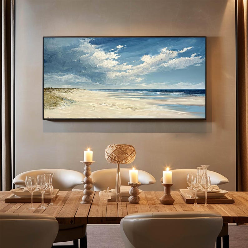 May include: A framed painting of a seascape with a sandy beach, blue sky, and white clouds. The painting is hanging on a wall above a wooden dining table with candles and a woven lampshade.