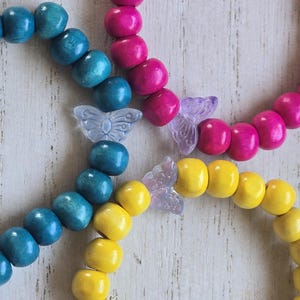 May include: Three beaded bracelets in teal, fuchsia, and yellow. Each bracelet is made of round wooden beads. Clear butterfly-shaped charms are interspersed between the beads. The bracelets are displayed on a white, textured surface.