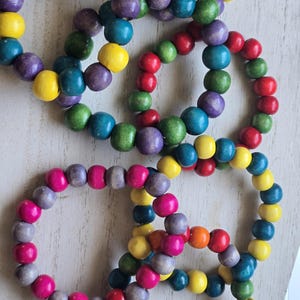 Op de afbeelding: Verschillende houten kralenarmbanden in diverse kleuren, waaronder geel, paars, blauw, groen, rood, roze en oranje. De armbanden zijn gemaakt van ronde kralen en liggen op een lichtgekleurd oppervlak.
