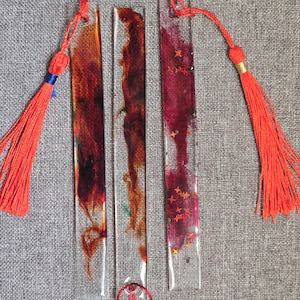 May include: Three rectangular resin bookmarks with colourful abstract designs. Each bookmark has a vibrant orange tassel attached. The designs include shades of red, orange, and pink, with some containing glitter.