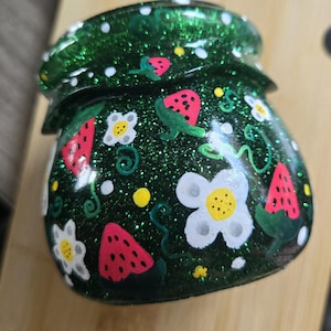 Puede incluir: Un tarro decorativo verde brillante con tapa, adornado con fresas pintadas a mano, flores blancas y enredaderas verdes. El tarro tiene un acabado brillante y está sobre una superficie marrón claro. Un artículo único y artesanal.