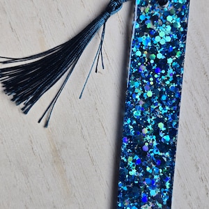 May include: A blue glitter bookmark with a dark blue tassel. The bookmark is rectangular and has a shiny, iridescent finish.