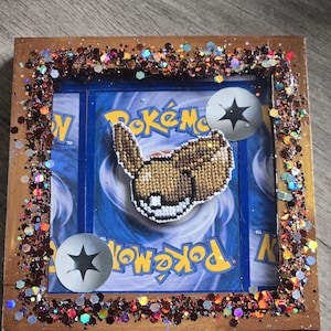 May include: A framed Pokémon card featuring a brown and white cross-stitch design of an Eevee character. The frame is decorated with glitter and two silver energy symbols. The background is a blue Pokémon card.