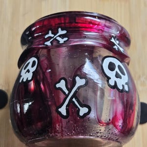 May include: A small, round, glass jar with a dark red and purple interior. The jar is decorated with white skull and crossbones designs. The jar has a wide opening and a slightly flared rim. The jar is likely used for storage or decoration.
