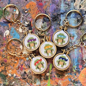 May include: Six colorful keychains featuring cross-stitched images of Teenage Mutant Ninja Turtles. Each keychain is attached to a gold-colored metal ring.