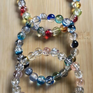 Può includere: Quattro braccialetti fatti di perline trasparenti e colorate. Le perline sono di vari colori, tra cui rosso, blu, giallo e verde. I braccialetti sono su una superficie di legno marrone chiaro.