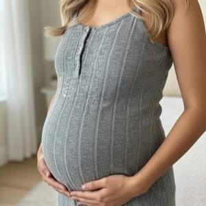 May include: A grey, ribbed maternity dress with a scoop neckline and button detail. The sleeveless dress is form-fitting and reaches the mid-thigh. The person is holding their pregnant belly. The background is a soft, neutral colour.