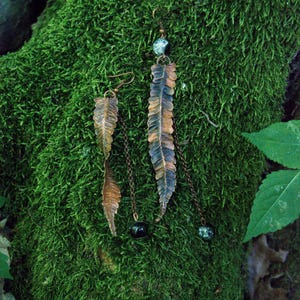 Puede incluir: Un par de pendientes colgantes con dijes de hoja de helecho en metal color cobre. Cada pendiente incluye una cadena metálica fina, una pequeña cuenta redonda con un patrón moteado en verde y negro, y un gancho para la oreja.